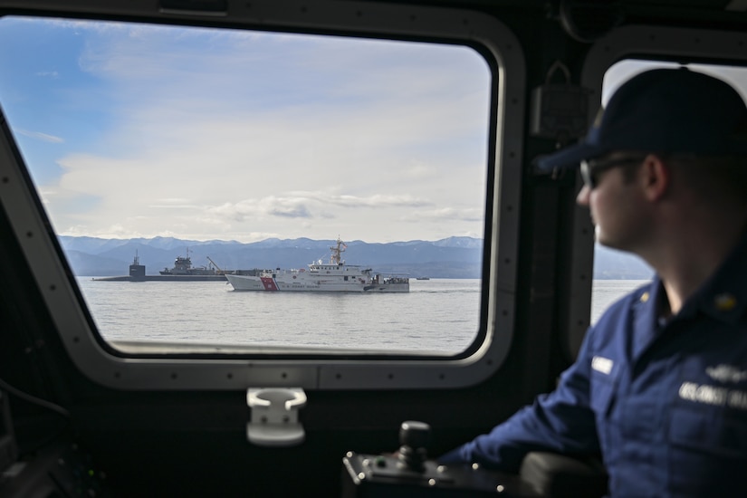 A man wearing a military uniform looks out the window of a boat at two other ships and a submarine sailing in the water with mountains in the background.