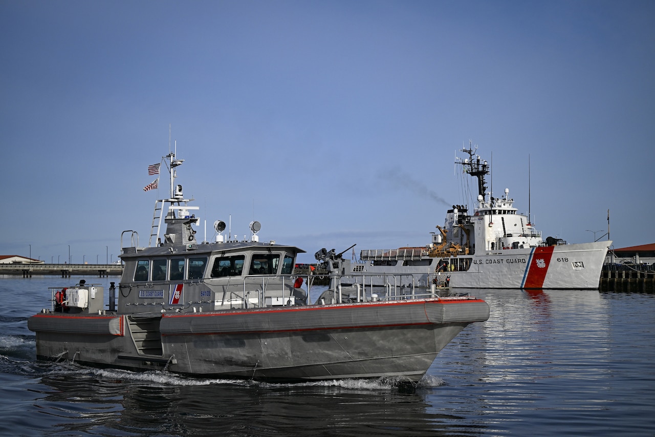 A medium-sized boat moves through the water as a larger ship is docked in the background.