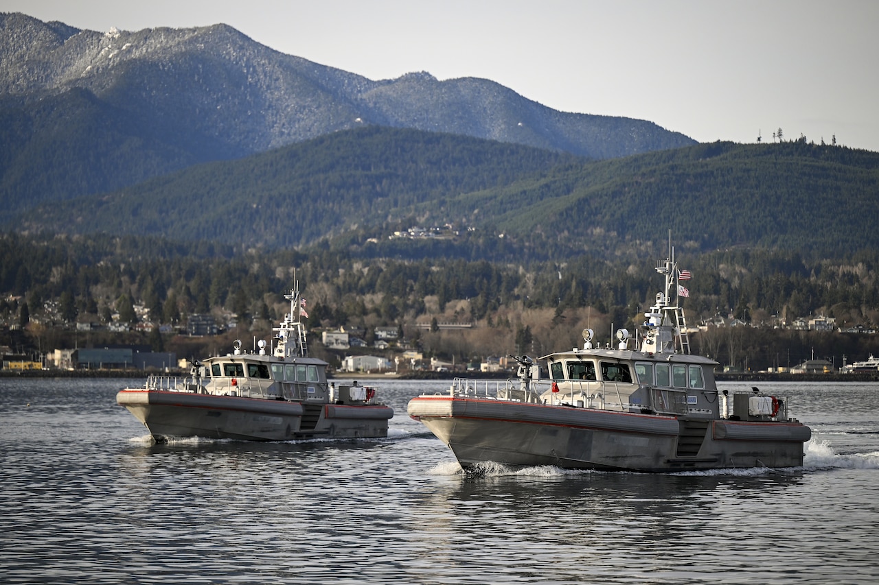 Two military boats sail in the water with houses and mountains in the background.