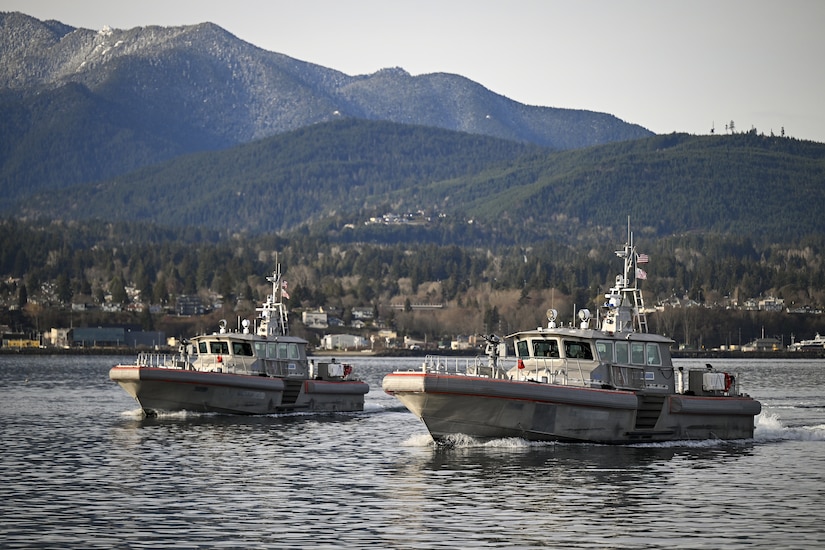 Two military boats sail in the water with houses and mountains in the background.