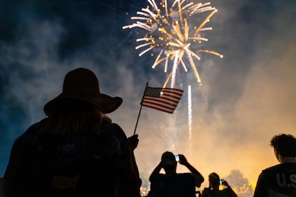 man with a hat holds a U.S. Flag as he watches the fireworks explode in the sky.