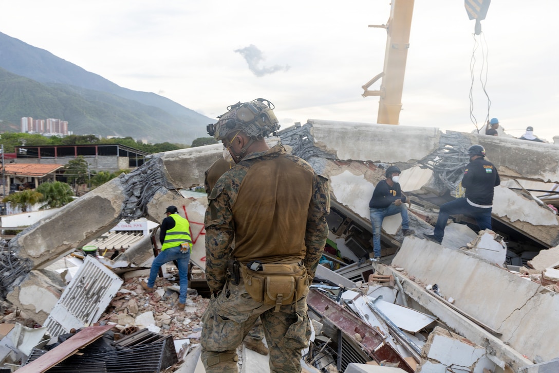 A Marine wearing a camouflage uniform, a helmet and protective face gear looks through large piles of debris with other people wearing civilian clothes during the day.