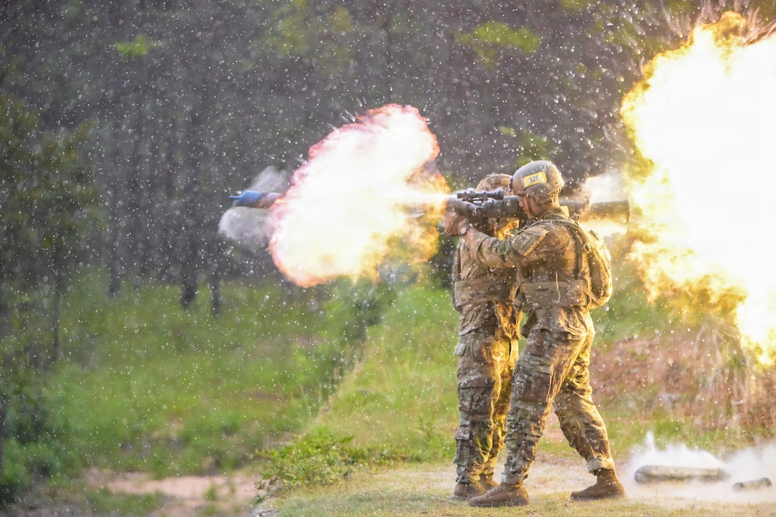Two Army trainees wearing camouflage uniforms, vests and helmets fire shoulder weapons in opposite directions creating orange fireballs and clouds of smoke in the woods as rain falls.