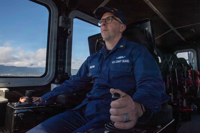 A coast guardsman in a blue uniform sits at the controls of a boat.