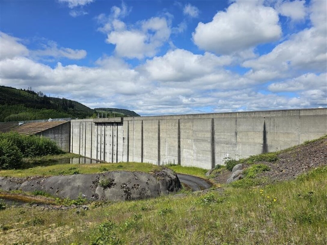 Under a blue sky with putffy white clouds, a tall cement wall stands between two grass-covered hills.
