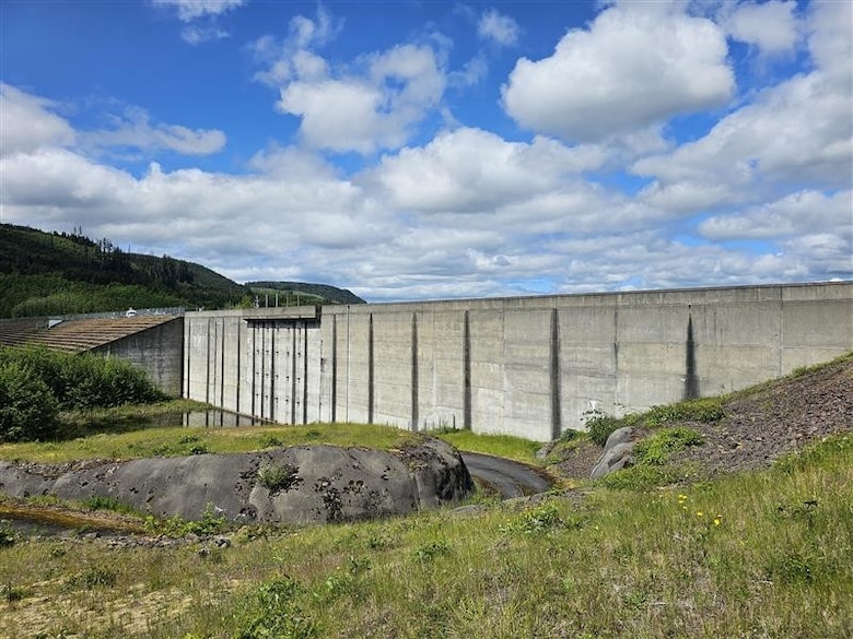 Under a blue sky with putffy white clouds, a tall cement wall stands between two grass-covered hills.