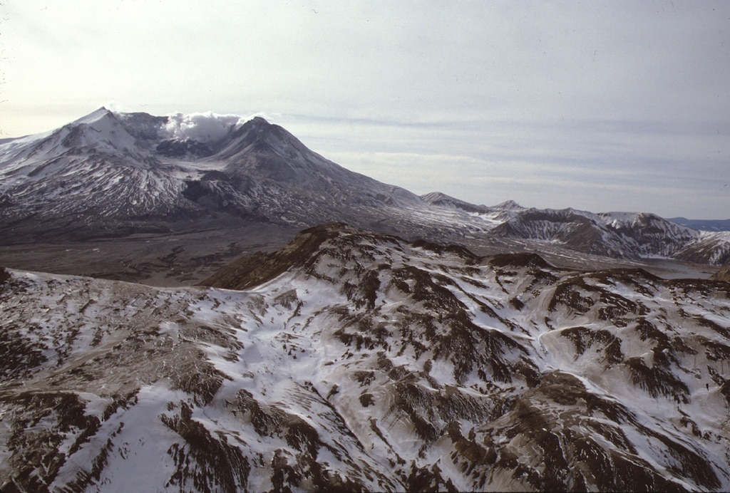 Under a cloudy sky, a rough landscape, lightly dusted with snow, is shown. The tips of the rough landscape are brown, the snow collects in the folds below, which is Mount St. Helen's pumice plain. In the background, Mount St. Helen looms, showing the collapsed top and side of the mountain from the 1980 explosion.
