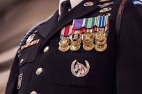 Only the chest is seen of an Army Soldier who is wearing a dark blue ceremonial uniform with many shiny medals pinned on one side and other ribbons and badges on other portions of the coat. Underneath the medals is a silver badge with a horse and horseshoe.
