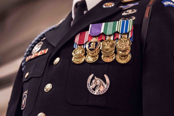 Only the chest is seen of an Army Soldier who is wearing a dark blue ceremonial uniform with many shiny medals pinned on one side and other ribbons and badges on other portions of the coat. Underneath the medals is a silver badge with a horse and horseshoe.