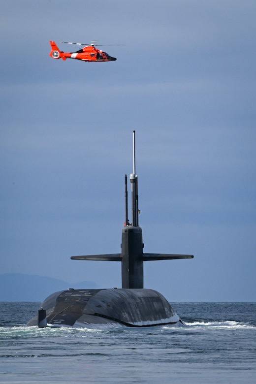 An orange helicopter flies high above a submarine making waves as it goes out to sea.
