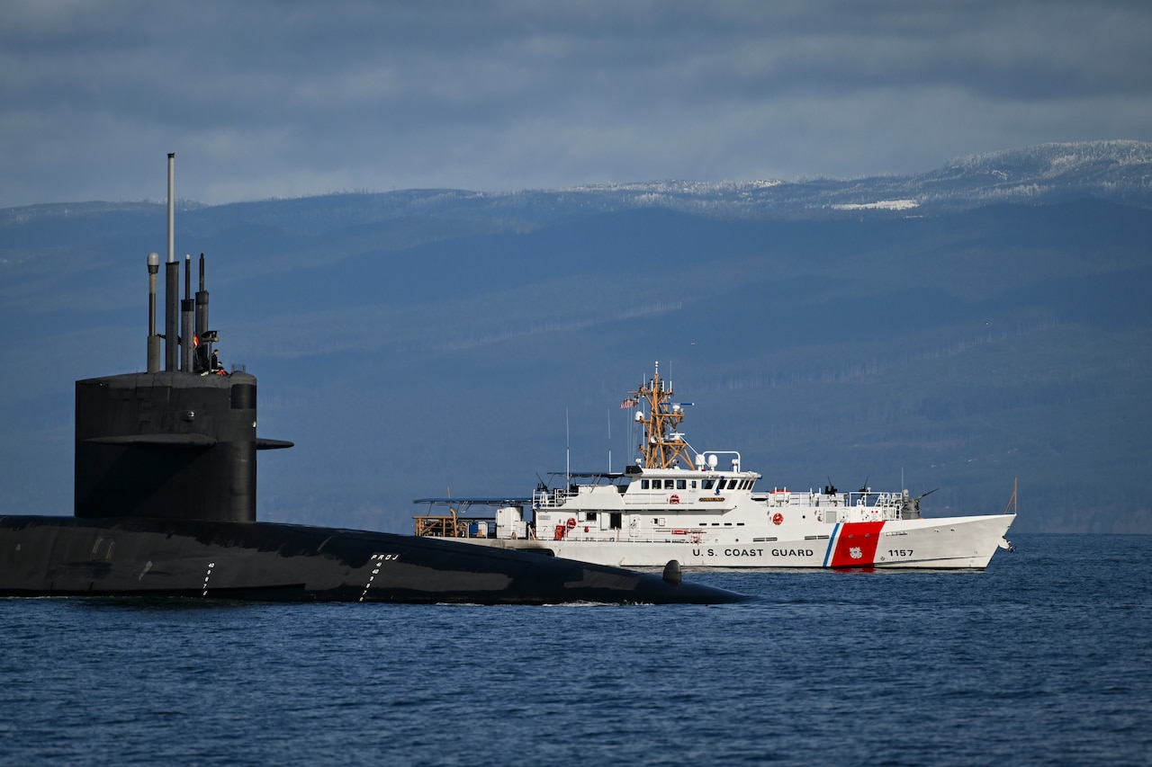 A Coast Guard ship sails next to a partially submerged submarine with mountains in the background.