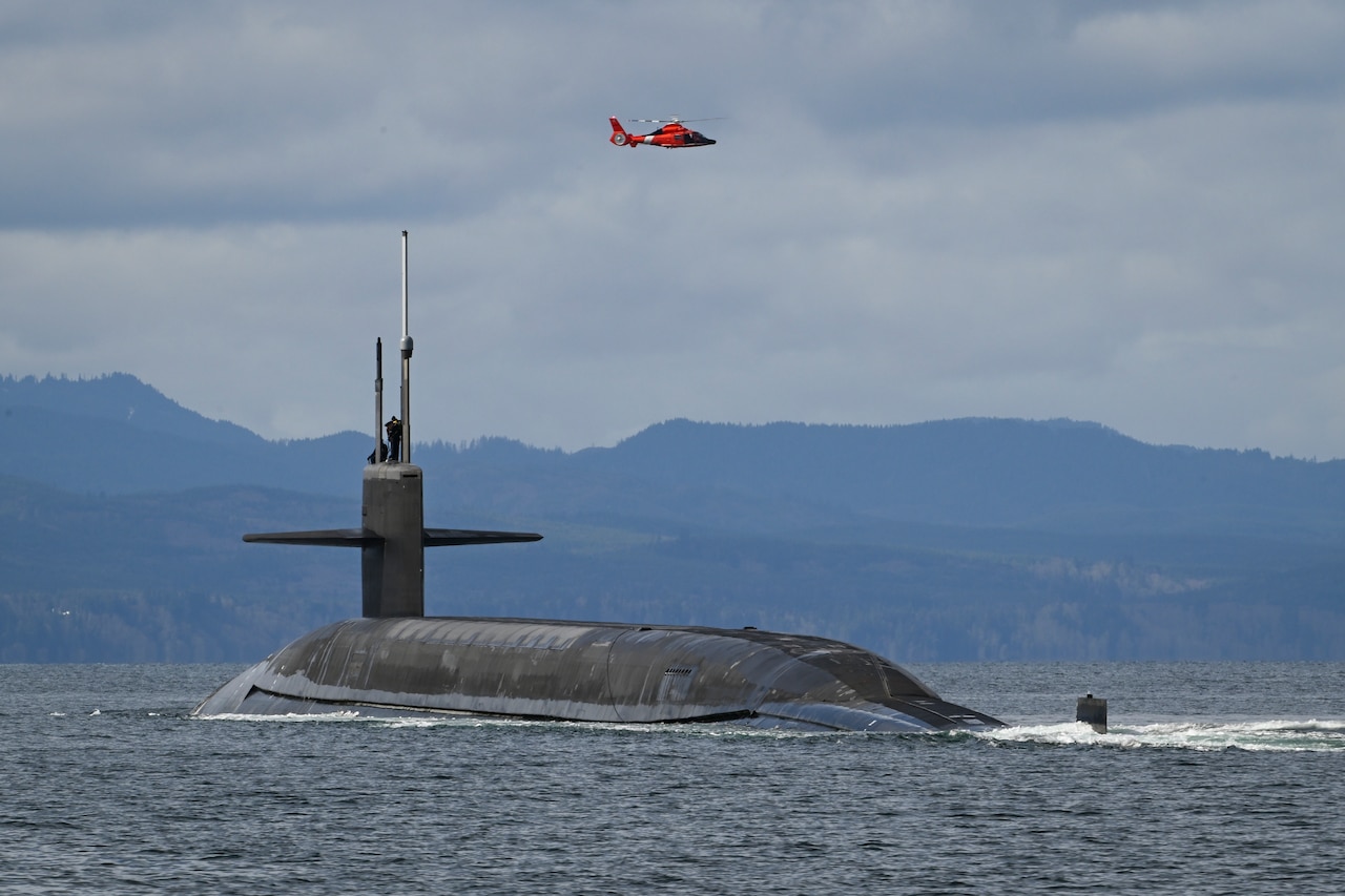 An orange helicopter flies high above a partially submerged submarine, with mountains in the distance.