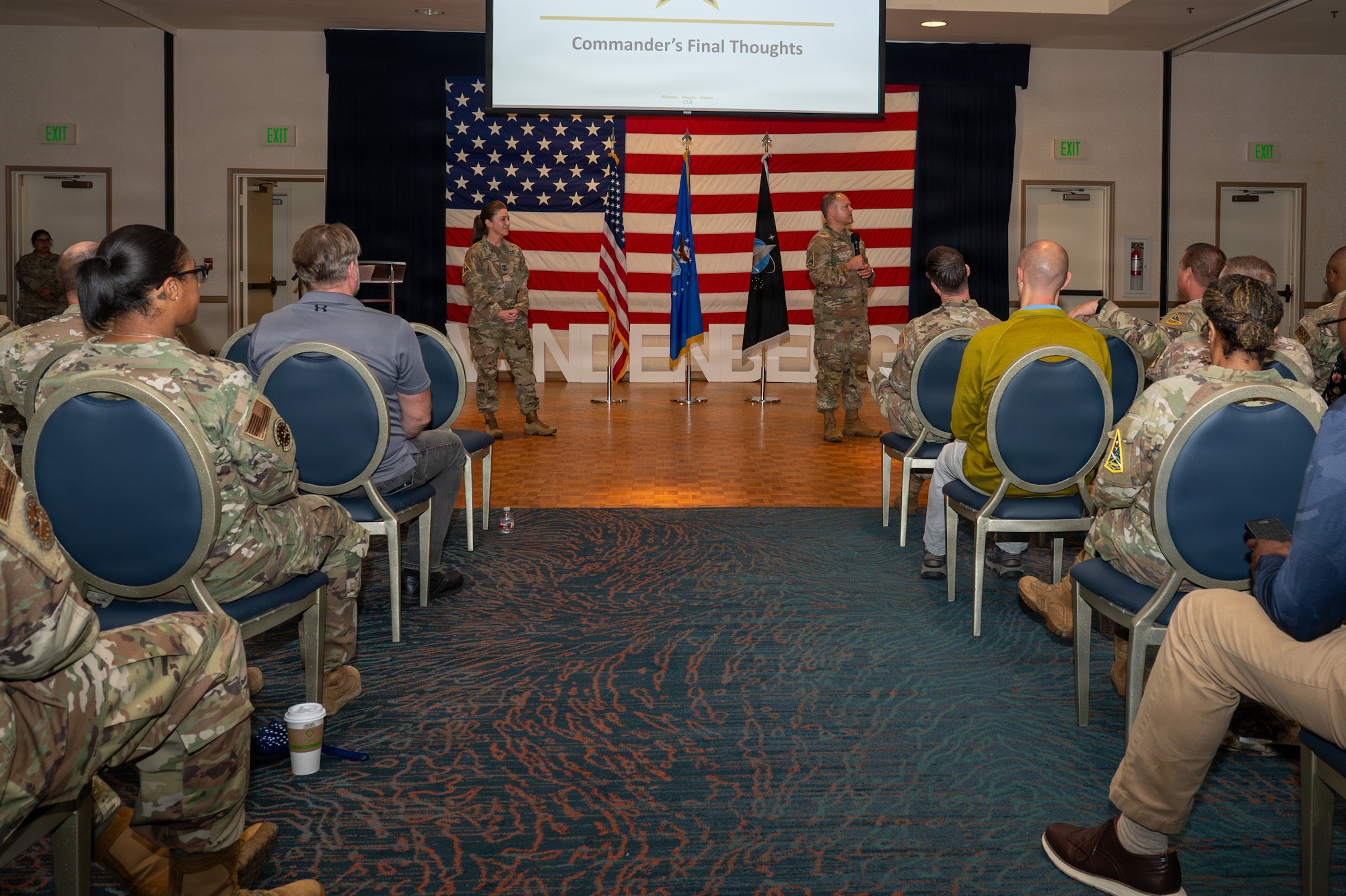 Two people stand on stage as others watch and listen