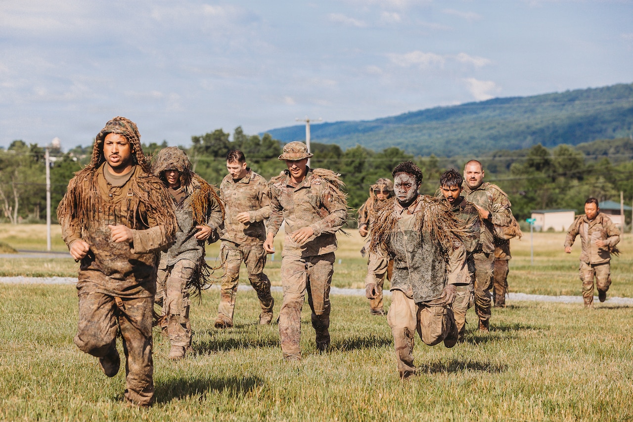 Nearly a dozen people in camouflage military uniforms and face paint jog through a grassy field with trees in the distance.