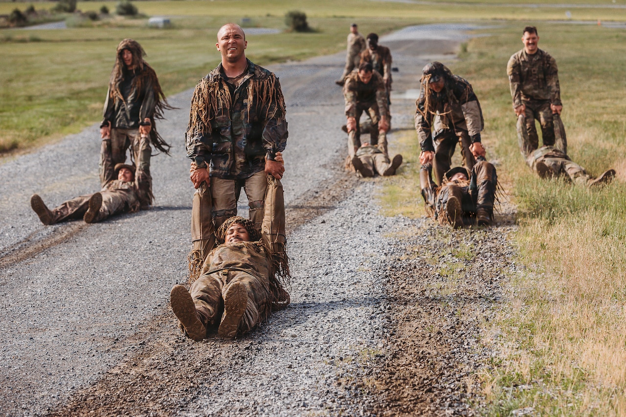 Five people in camouflage military uniforms each drag a person in similar attire down a gravel road.