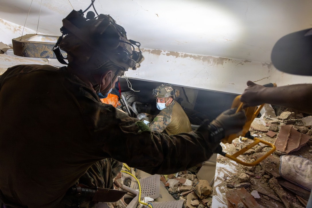Two service members wearing military uniforms and helmets work in rubble and debris with a third partly visible person.