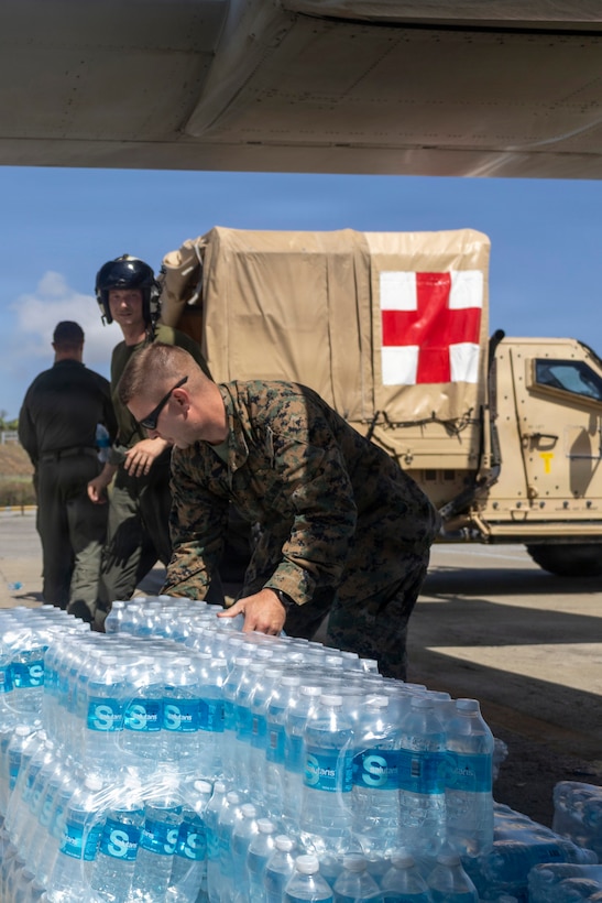 Three Marines work on a flight line, one handling cases of water and two others near a military ambulance.