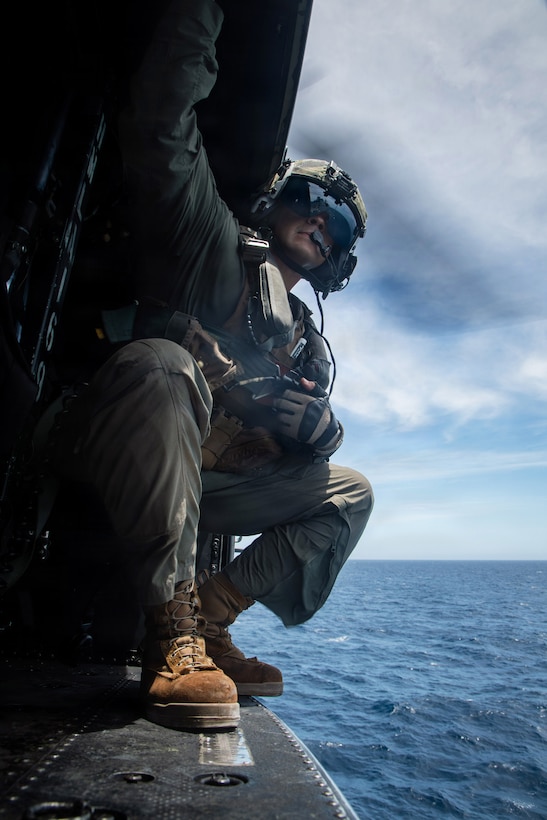 A Marine looks out from the open door of a helicopter in flight over water.
