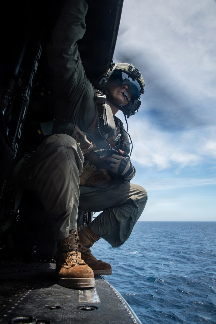 A Marine looks out from the open door of a helicopter in flight over water.