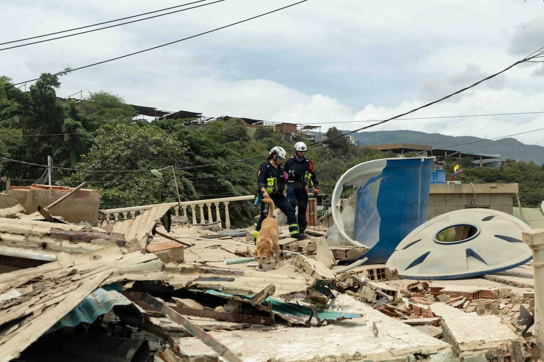 Two civilians in rescue gear walk with a sniffing dog through building wreckage.