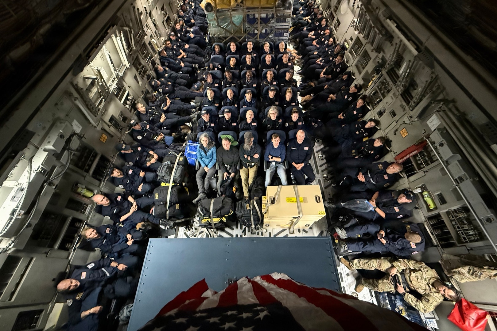 Civilian rescue personnel sit aboard a packed aircraft, with a U.S. flag hanging above them.