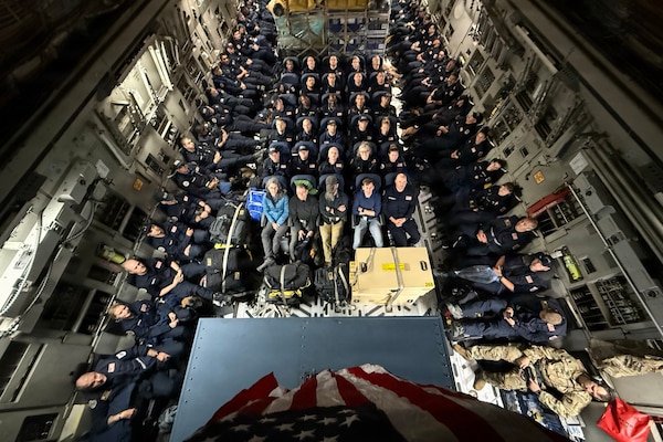 Civilian rescue personnel sit aboard a packed aircraft, with a U.S. flag hanging above them.