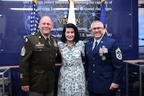 Two people in military uniforms flank a person in civilian clothing while standing in front of flags.