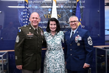 Two people in military uniforms flank a person in civilian clothing while standing in front of flags.