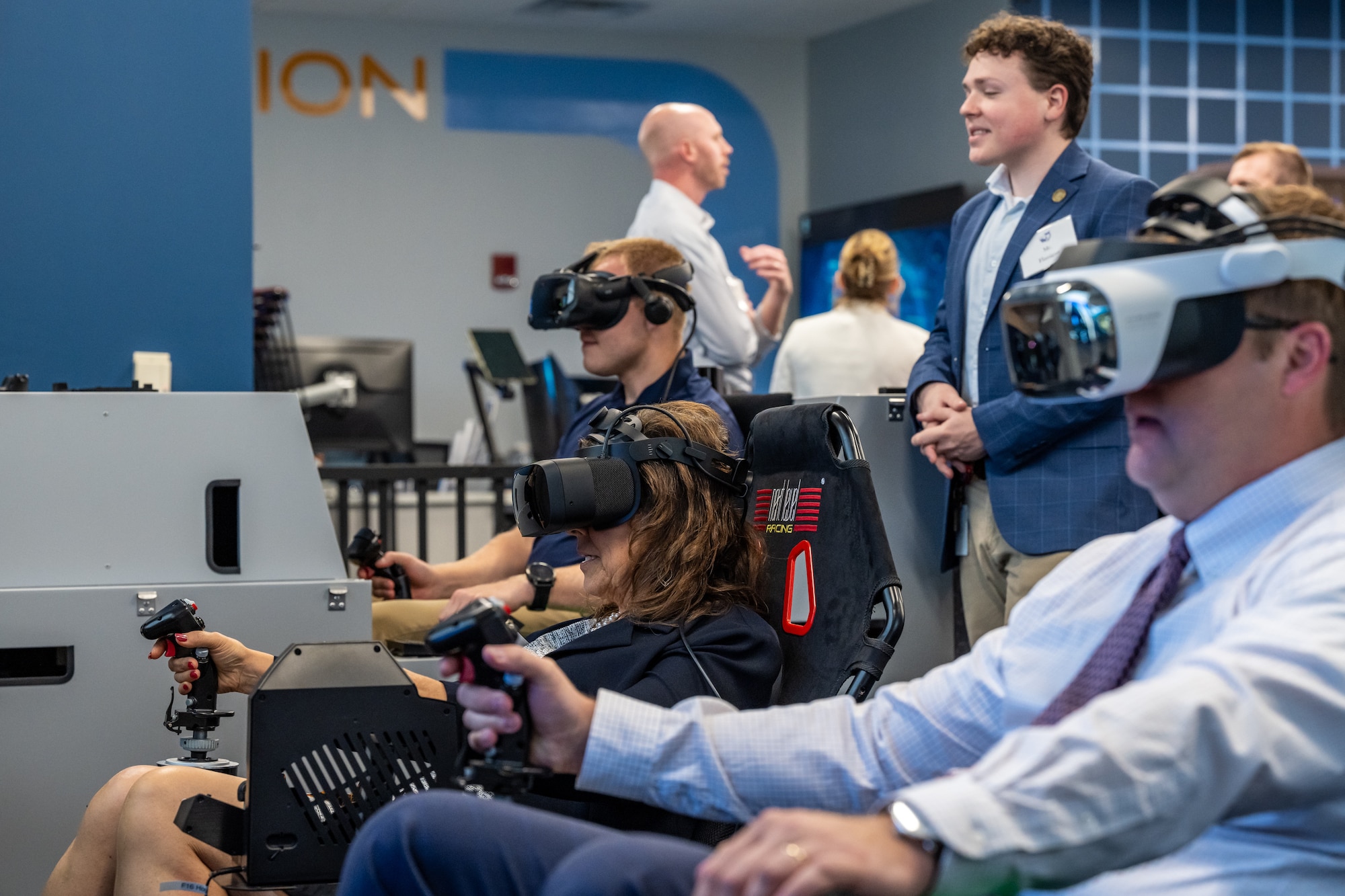Men and women, wearing business casual attire, sit in flight simulator chairs with virtual reality headsets on