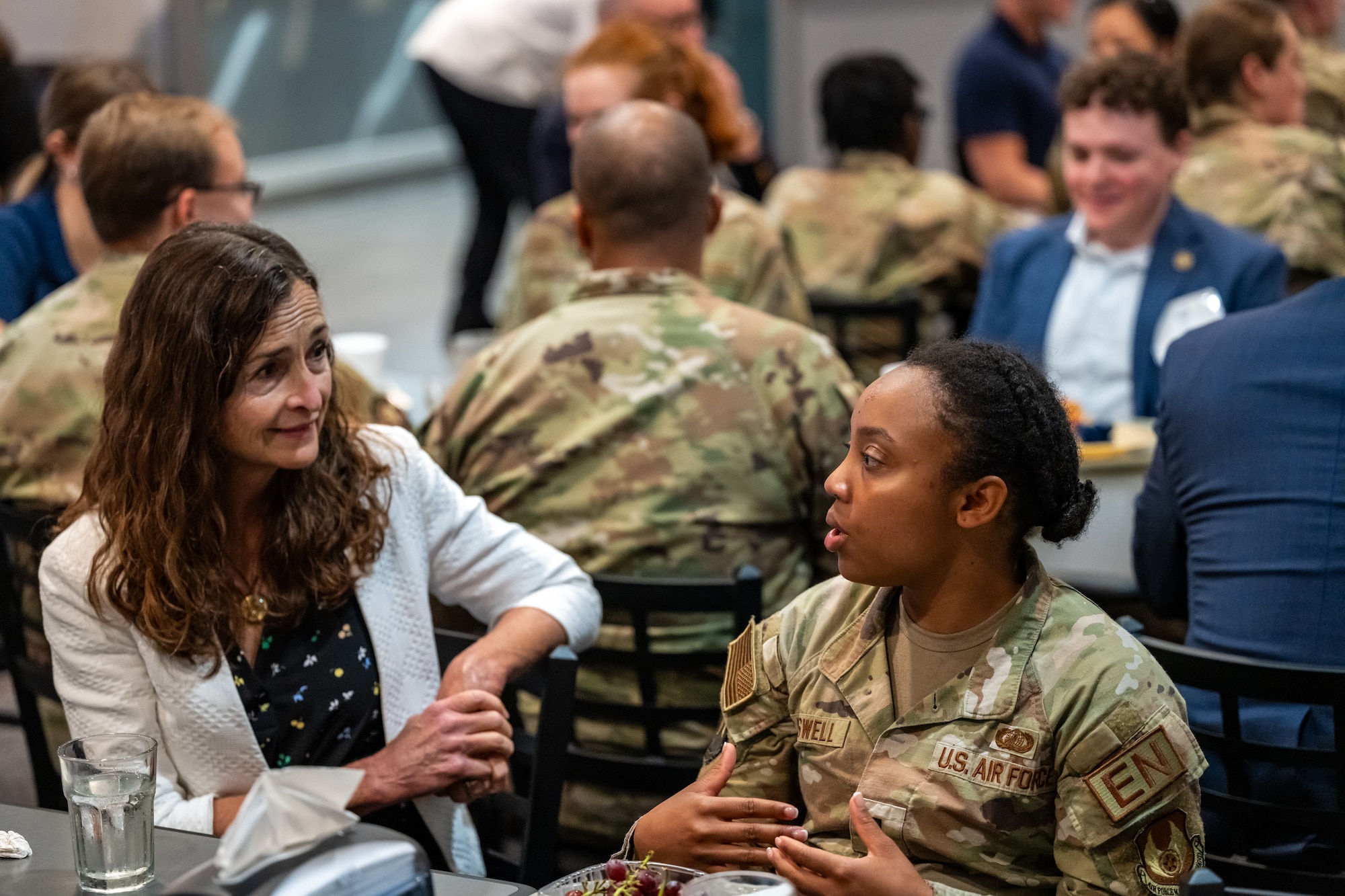 Two women, one wearing a white blazer one wearing a military camouflage uniform, talk with one another, more civilians and military members can be seen sitting in background