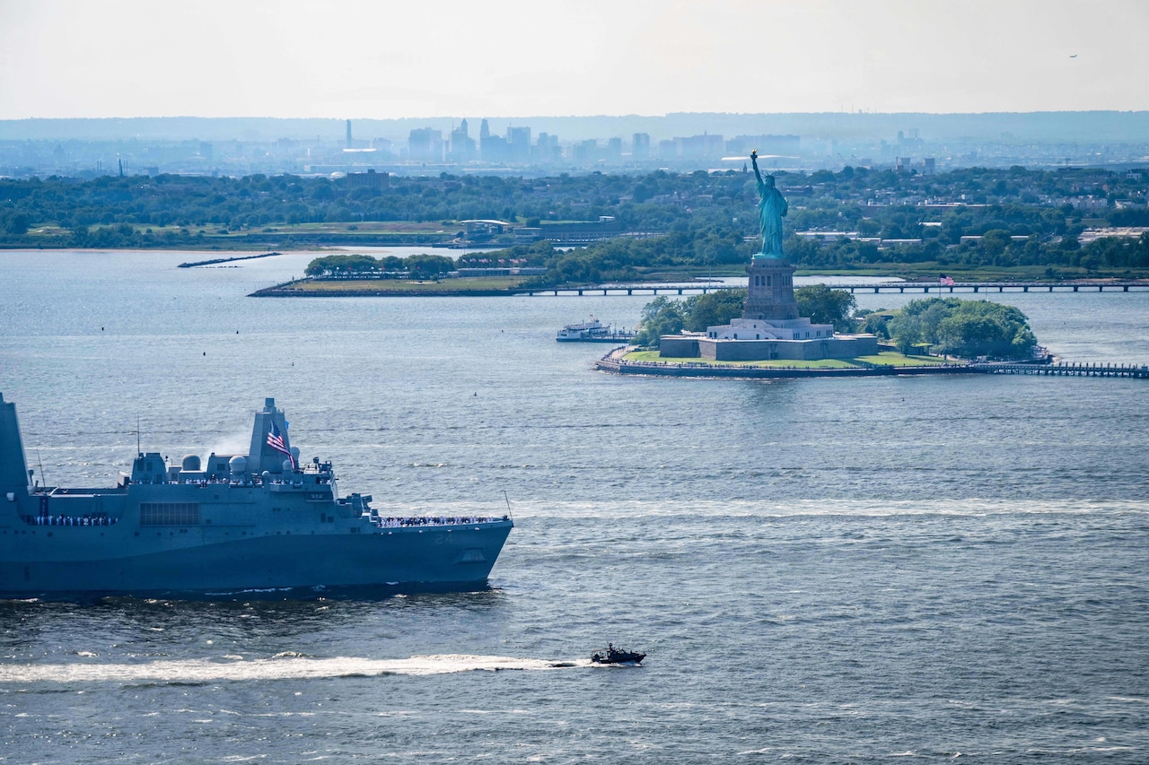A Navy ship and small boat transit a body of water near the Statue of Liberty with the New York skyline in the distance.
