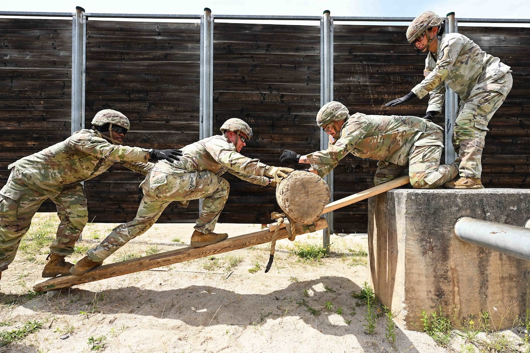 Four soldiers in camouflage uniforms roll a log up a diagonal wooden plank to a concrete cube in the sand in front of a fence.