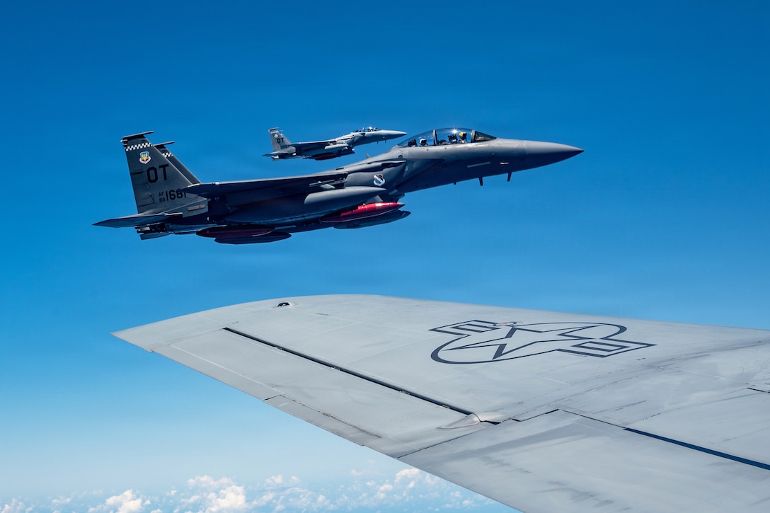 Two aircraft fly in formation in a blue sky as seen over the wing of another aircraft in the foreground.