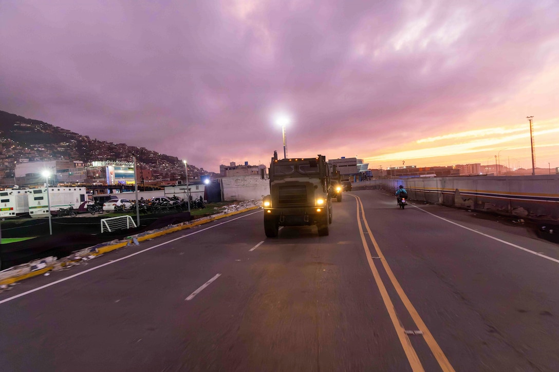 Military trucks drive on the road under a purplish, golden sky with homes on a mountain to the left and a motorist going in the opposite direction on the right.