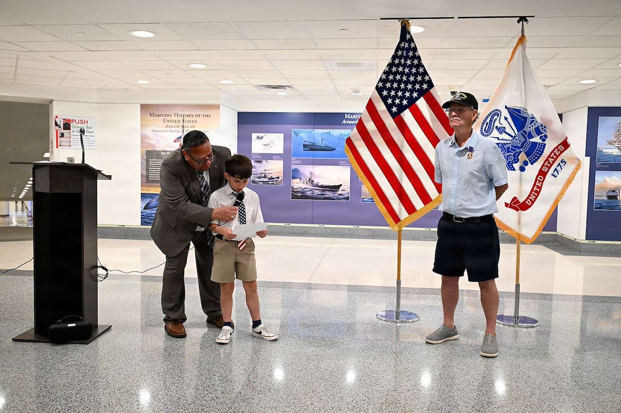 A man in business attire crouches to hold a microphone for a boy who is speaking into a microphone while another man stands to the side at parade rest. An American flag and an Army flag are in the background.
