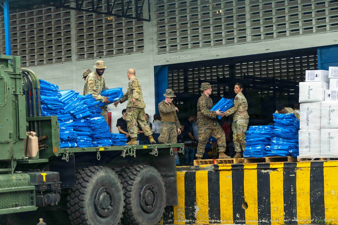 Soldiers and Marines help each other load blue, folded material onto the back of a vehicle with a flat bed.