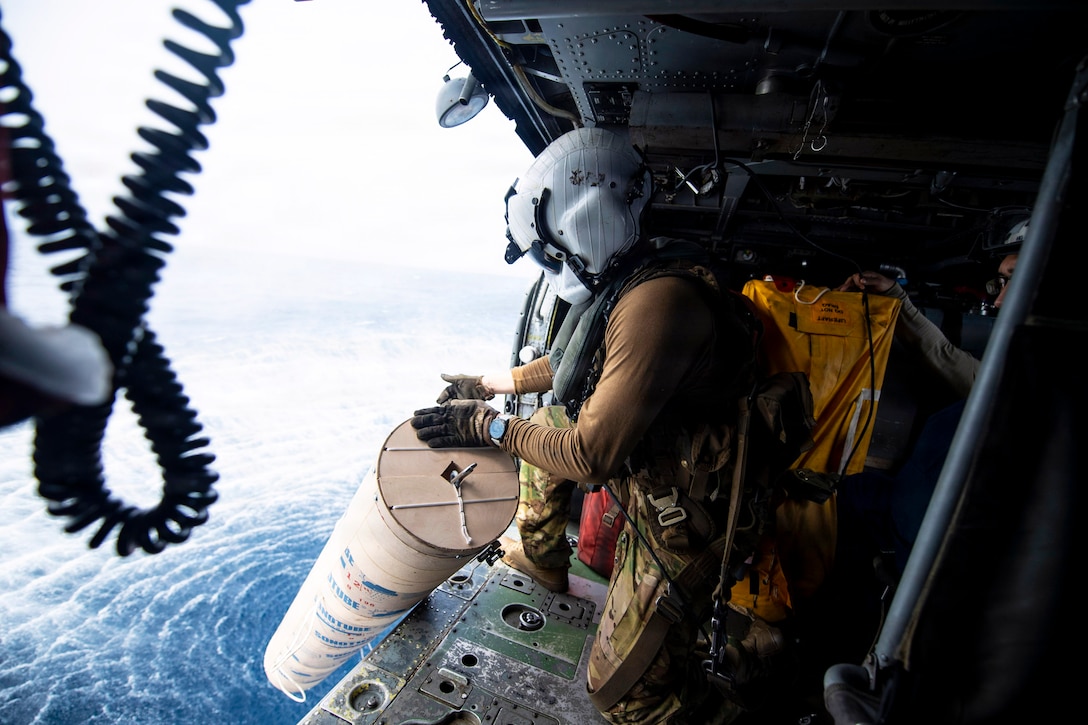 A sailor whose entire face is covered by a white helmet and dark glasses sits in a helicopter cabin over the ocean and drops a device into it.