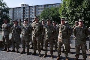 A group of men wearing camouflage military uniforms are standing outside in ranks while holding their hands behind their backs.