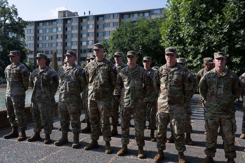 A group of men wearing camouflage military uniforms are standing outside in ranks while holding their hands behind their backs.