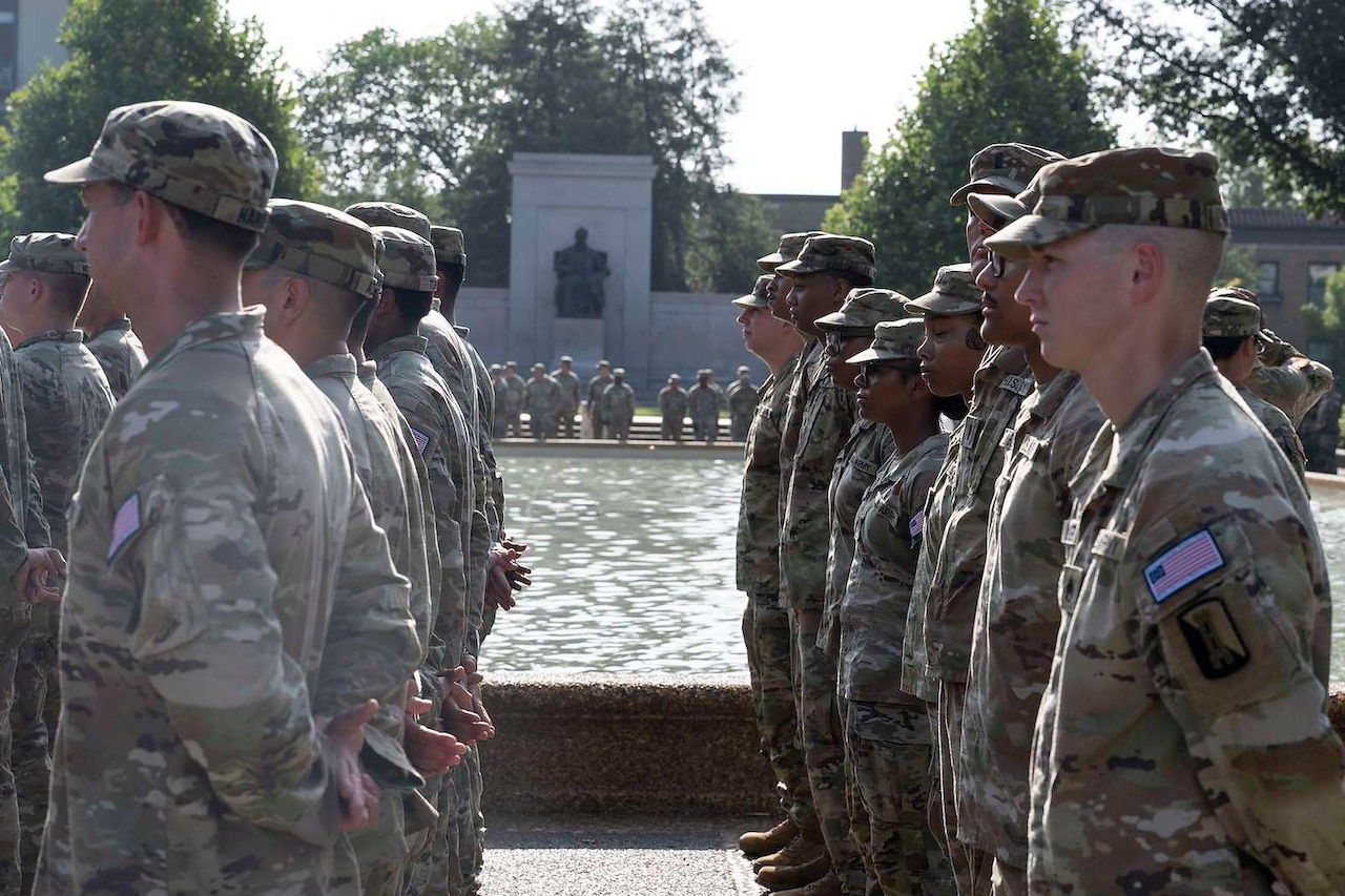 A group of men and women wearing camouflage military uniforms are standing outside in ranks while holding their hands behind their backs. A large fountain and additional people in uniform are behind them.