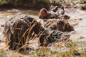 Four people in camouflage military uniforms crawl through a muddy stream on their stomachs.