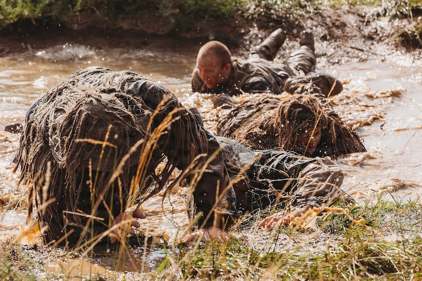 Four people in camouflage military uniforms crawl through a muddy stream on their stomachs.