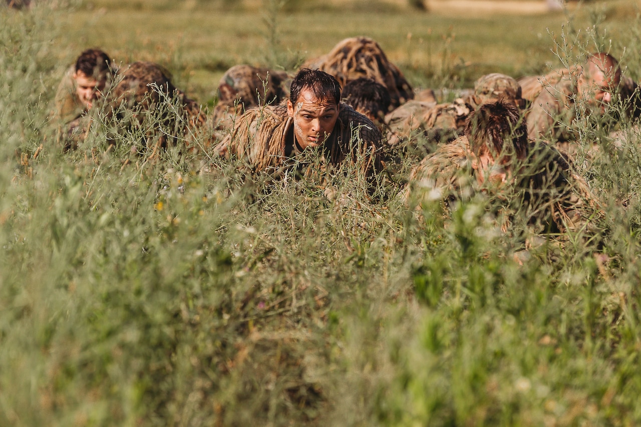About six people in camouflage military uniforms and face paint crawl through tall grass.