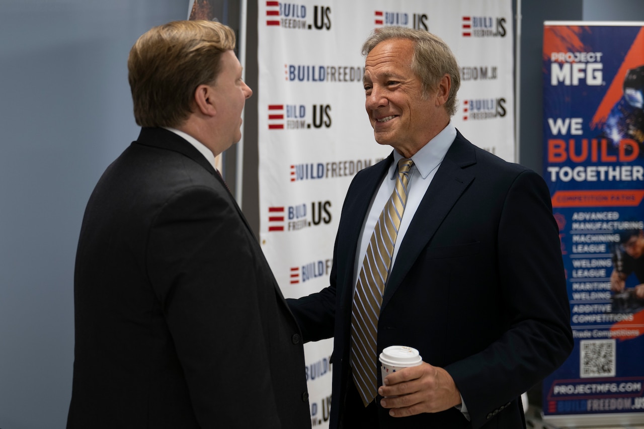 Two men in business attire stand and speak to each other indoors; one is holding a cup.