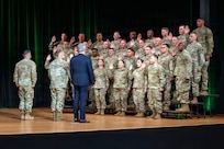 Dozens of Army Soldiers wearing combat uniforms are standing on risers on a stage and raising there right hands while three people, two in combat uniforms and one in a business suit, face them during a ceremony.