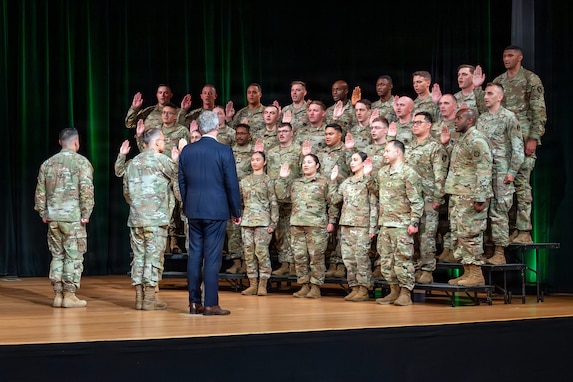 Dozens of Army Soldiers wearing combat uniforms are standing on risers on a stage and raising there right hands while three people, two in combat uniforms and one in a business suit, face them during a ceremony.