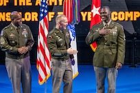 An Army Soldier wearing a green service dress uniform on the right has his hand on his chest as two other similarly dressed Soldiers look on. They are standing in front of flags during an indoor ceremony.