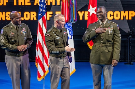 An Army Soldier wearing a green service dress uniform on the right has his hand on his chest as two other similarly dressed Soldiers look on. They are standing in front of flags during an indoor ceremony.