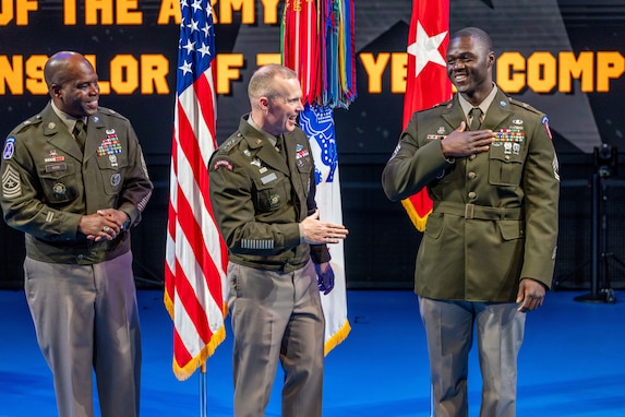 An Army Soldier wearing a green service dress uniform on the right has his hand on his chest as two other similarly dressed Soldiers look on. They are standing in front of flags during an indoor ceremony.