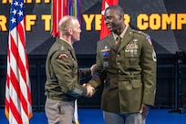 An Army Soldier wearing green service dress uniform is shaking hands with another similarly dressed Soldier while standing in front of flags during a ceremony.